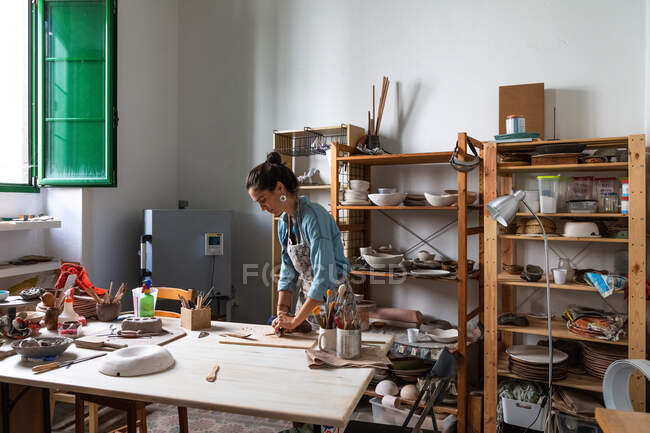 Señora adulta enfocada en ropa casual y delantal esculpir pieza de arcilla y sonreír durante la clase de cerámica en el taller - foto de stock