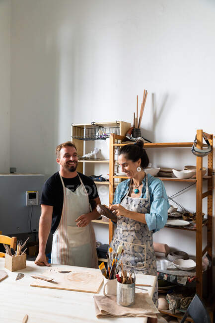 Positive middle aged female and young male in casual clothes and aprons smiling while creating clay plate during pottery class in workshop — Stock Photo