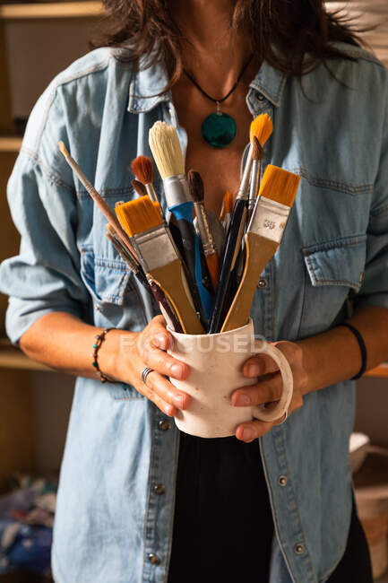 Cropped unrecognizable female master in casual clothes smiling and looking at camera while standing near shelf in workshop with mug of various paintbrushes in hand — Stock Photo