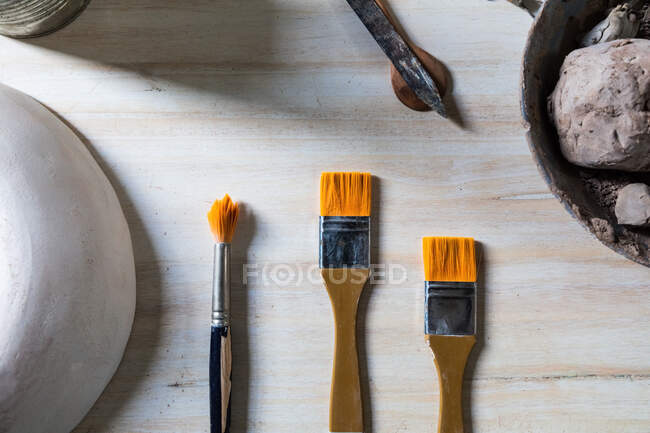 Набор керамики и кистей для рисования на деревянном столе с тарелкой глины и керамическим горшком в мастерской — стоковое фото