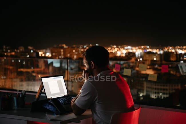 Back view of thoughtful young male freelancer in casual outfit sitting on modern building rooftop and working remotely on laptop in evening — Stock Photo
