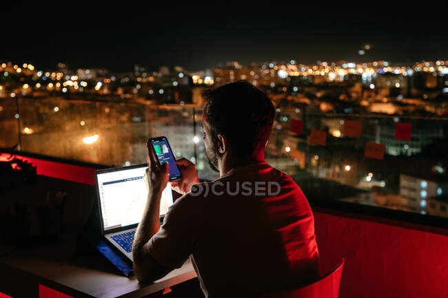 Вид ззаду на невизначений концентрований молодий чоловічий фрілансер у випадкових обмінних повідомленнях на смартфоні, сидячи на сучасному будівельному даху та працюючи віддалено на ноутбуці ввечері — стокове фото