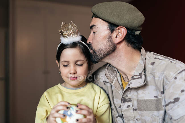 Бородатый мужчина в военной форме целует маленькую девочку, сидя рядом, прежде чем отправиться служить стране. — стоковое фото