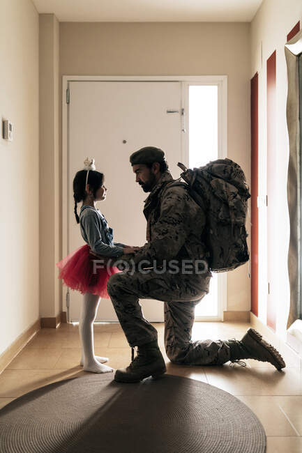 Вид сбоку полное тело солдата в камуфляжной одежде, стоящего на коленях перед военной службой с маленькой девочкой — стоковое фото