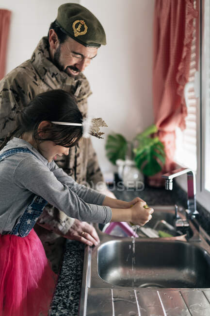 Вид збоку веселого військового чоловіка в уніформі, що стоїть біля раковини з чарівною дочкою і миє посуд разом на кухні вдома — стокове фото