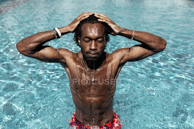Homem afro-americano despreocupado de pé com os olhos fechados na piscina e mãos na cabeça, desfrutando de férias de verão. — Fotografia de Stock