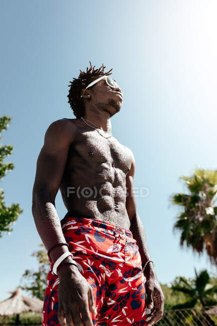 Уверенный молодой афроамериканец в обнаженном туловище в солнцезащитных очках и глядя вверх, стоя у бассейна после купания — стоковое фото