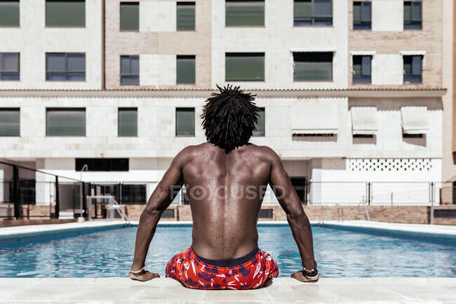 Vista trasera de un irreconocible hombre afroamericano sin camisa sentado junto a la piscina y disfrutando de un día soleado durante las vacaciones de verano - foto de stock