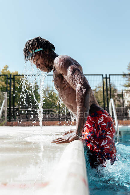 Seitenansicht des nassen afroamerikanischen Männchens, das aus einem Swimmingpool auftaucht und Wasser spritzt — Stockfoto