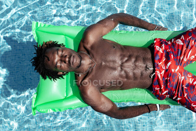 From above of carefree African American male with naked torso and in shorts lying eyes closed on inflatable mattress in swimming pool and enjoying sunny day during summer holiday — Stock Photo