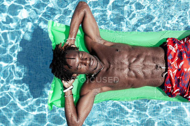 De cima de homem afro-americano despreocupado com tronco nu e calções deitado no colchão inflável na piscina e desfrutando de dia ensolarado durante as férias de verão — Fotografia de Stock