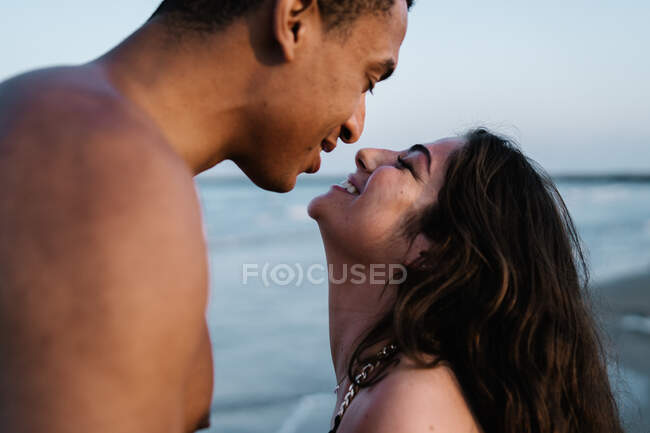 Viajero masculino afroamericano besando a su pareja femenina sincera de pie contra el océano durante el viaje de verano - foto de stock