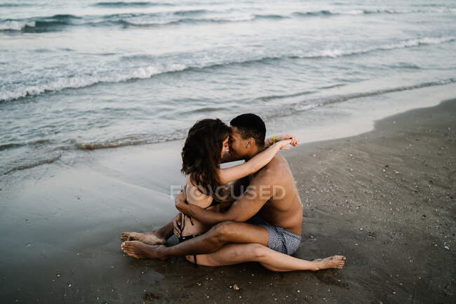 Вид збоку на молодий зміст босоніж багаторасової пари, що обіймається на піщаному океані пляжу під час літньої подорожі — стокове фото