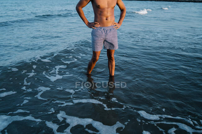 Cropped unrecognizable shirtless African American male traveler in shorts standing with hands on waist in foamy ocean — Stock Photo