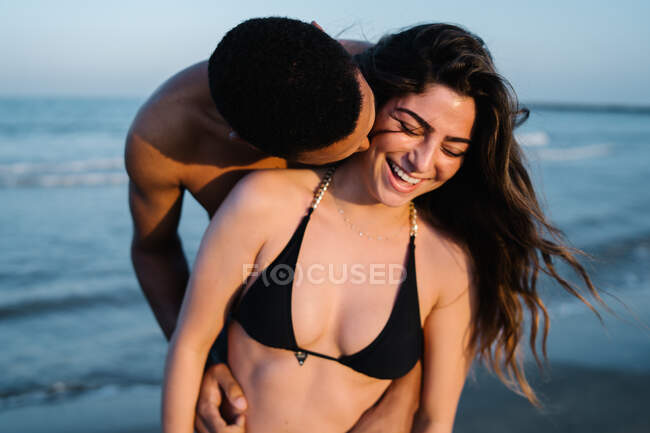 Anonymous African American male traveler kissing sincere female partner on cheek against ocean during summer trip — Stock Photo
