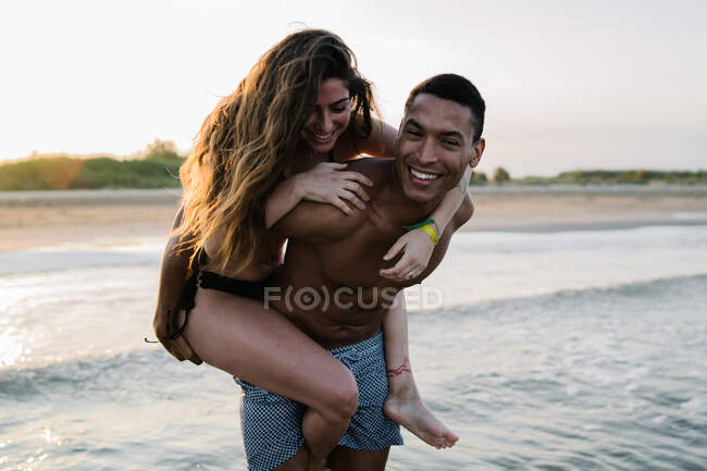 Joven mujer feliz turista cabalgando a cuestas en novio afroamericano contra el océano ondulado durante la luna de miel - foto de stock
