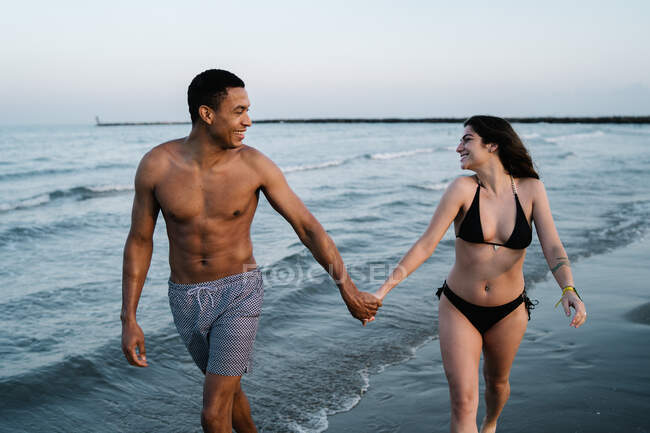 Zufriedene junge multiethnische Paar von Touristen in Badeanzügen schlendern an der Küste des Ozeans, während sie einander im Sommer betrachten — Stockfoto