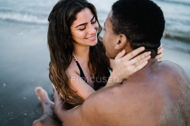 Молодий зміст босоніж багаторасової пари обіймається на піщаному океані пляжі під час літньої подорожі, дивлячись один на одного — стокове фото