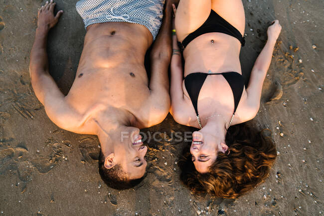 Top view of young content multiracial couple of travelers in swimwear looking at each other while lying on coast — Stock Photo