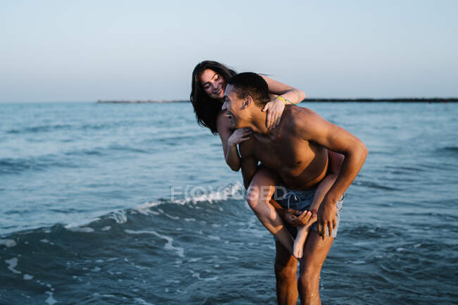 Young happy female tourist riding piggyback on African American boyfriend against wavy ocean during honeymoon — Stock Photo