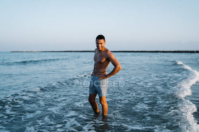 Young cheerful shirtless African American male traveler in shorts standing with hands on waist in foamy ocean — Stock Photo