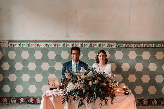 Молода багаторасова наречена і наречена сидять за столом з квітковим букетом і полум'яними свічками, дивлячись на камеру в ресторані — стокове фото