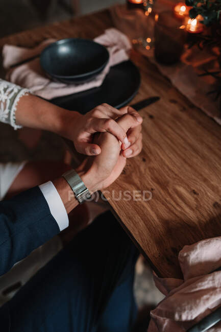 Crop anonymous groom and bride holding hands on served table with flower decor and burning candles in restaurant — Stock Photo