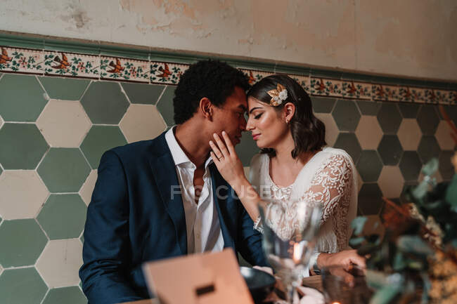 Jovem noiva multirracial e noivo acariciando uns aos outros enquanto se sentam à mesa com buquê de flores e velas flamejantes no restaurante com os olhos fechados — Fotografia de Stock