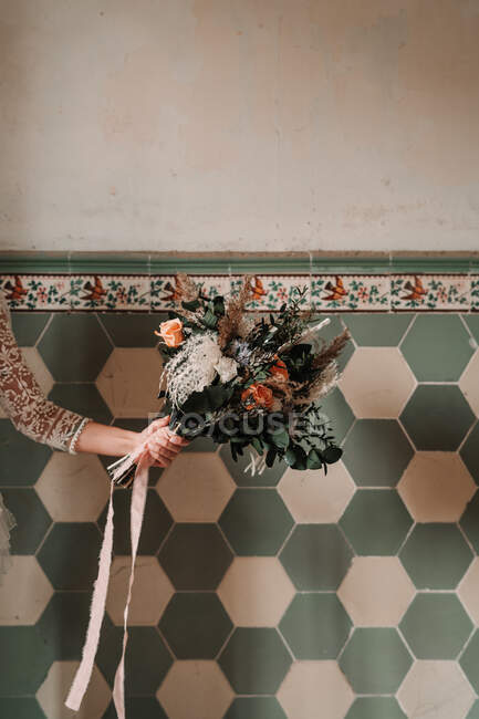 Crop anonymous woman with blooming flowers and ribbon near tiled wall with decor on wedding day in building — Stock Photo