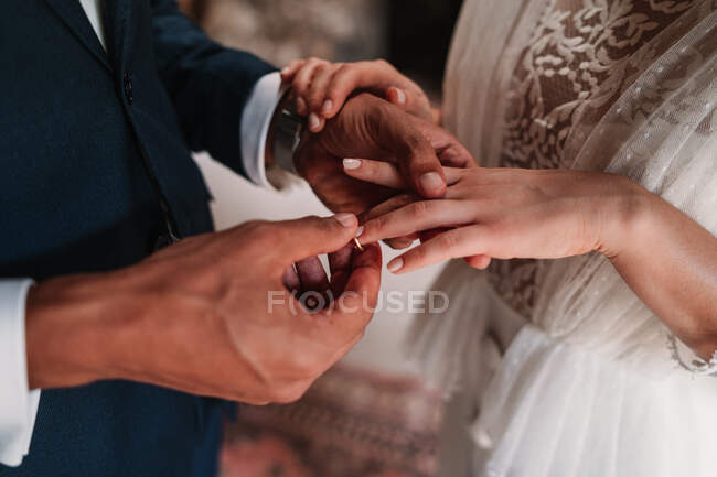 Высокоугольный вид сбоку обрезанный неузнаваемый этнический жених надевает кольцо на палец невесты в модных свадебных платьях, нежно держась за руки с любовью — стоковое фото