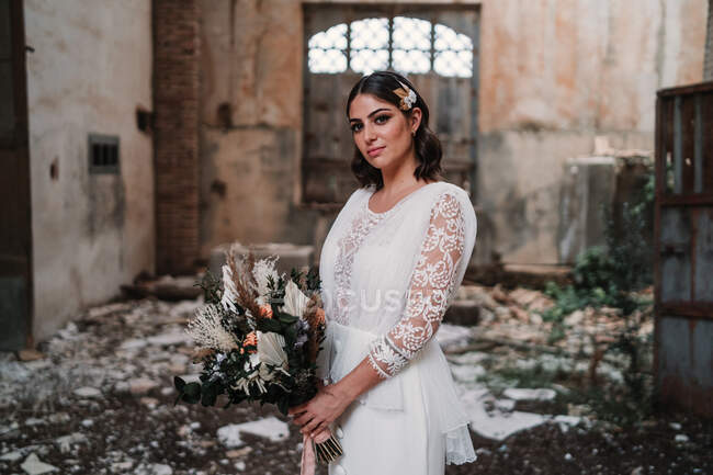 Heitere junge Braut in elegantem weißen Kleid mit zartem Strauß steht in verlassenen Ruinen und blickt in die Kamera — Stockfoto