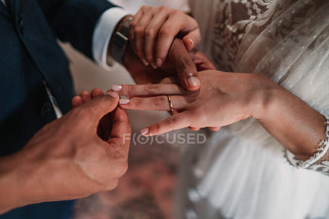 Vue latérale à angle élevé de marié ethnique méconnaissable recadrée mettant anneau sur doigt de la mariée dans des robes de mariée fantaisie tenant la main doucement avec affection — Photo de stock