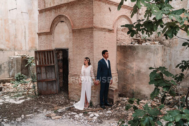 Cuerpo completo joven pareja étnica usando vestidos de novia elegantes tomados de la mano fuera abandonado edificio en ruinas - foto de stock