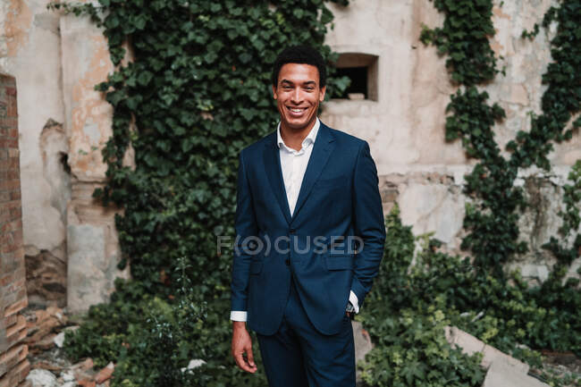 Happy ethnic male in elegant suit standing with hand in pocket and looking at camera against weathered building wall — Stock Photo
