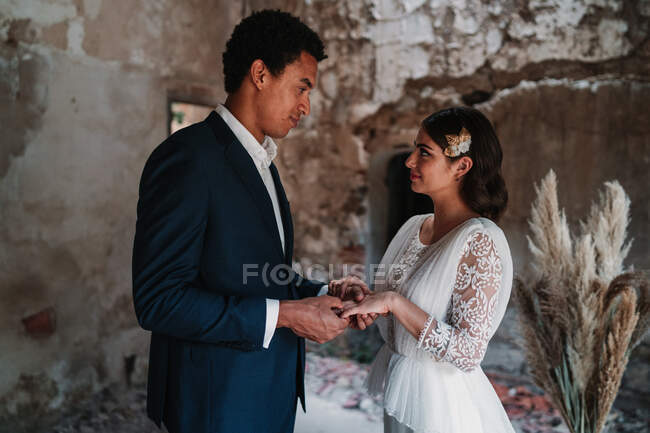 Side view content young ethnic groom putting ring on finger of bride in fancy wedding gowns holding hands gently and looking at each other with affection while standing in grunge cracked building — Stock Photo