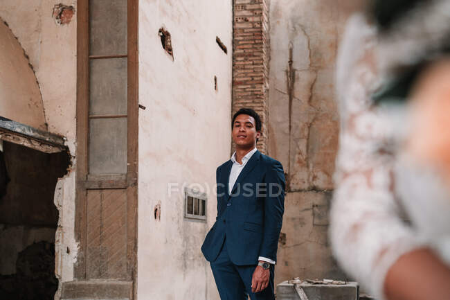 Content young ethnic groom wearing classy suit standing looking at camera near cropped unrecognizable bride in obsolete ruined building room — Stock Photo