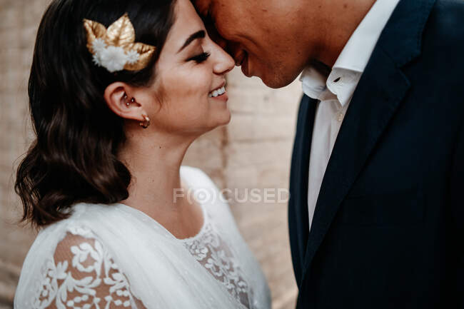 Side view romantic ethnic newlywed couple in elegant clothes bonding tenderly with eyes closed in light wedding studio — Stock Photo