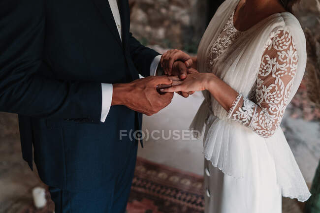 Вид сбоку обрезанный неузнаваемый этнический жених надевающий кольцо на палец невесты в модных свадебных платьях, нежно держащийся за руки с любовью — стоковое фото