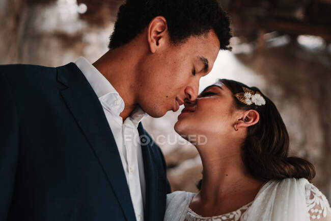 Vista lateral romântico casal recém-casado étnico em roupas elegantes ligando ternamente com os olhos fechados no estúdio de casamento leve — Fotografia de Stock