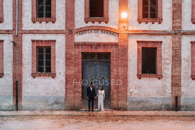 Full length loving ethnic couple wearing elegant wedding clothes holding hands and looking at each other while standing outside aged brick building — Stock Photo