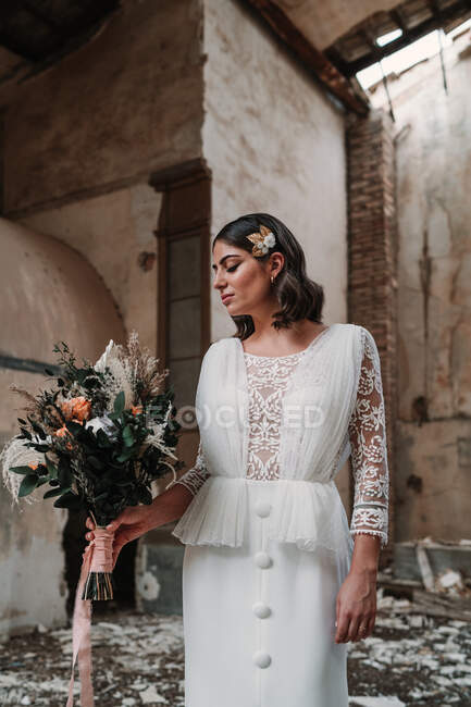Serene young bride wearing elegant white dress with delicate bouquet standing in abandoned ruined building with eyes closed — Stock Photo