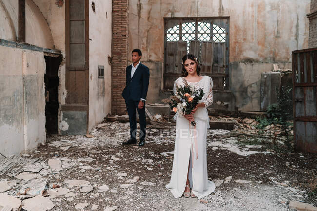 Content young bride wearing classy wedding gown with tender bouquet standing looking at camera near ethnic groom in obsolete ruined building room — Stock Photo