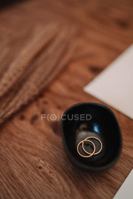 De arriba anillos de boda clásicos de oro en cuenco negro colocado en la mesa de madera en el estudio de la boda - foto de stock