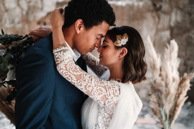 Вид сбоку романтическая этническая новобрачная пара в элегантной одежде нежно сближаясь с закрытыми глазами в светлой свадебной студии — стоковое фото