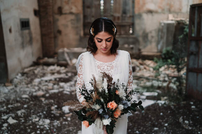 Спокойная молодая невеста в элегантном белом платье с тонким букетом стоит в заброшенном разрушенном здании с закрытыми глазами — стоковое фото