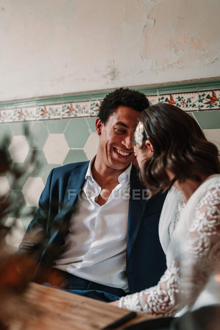 Cheerful young multiracial bride and groom sitting at table with flower bouquet and flaming candles while looking at each other in restaurant — Stock Photo