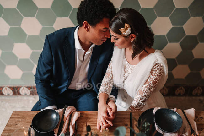 Зверху молода багаторасова наречена і наречена пестить один одного, сидячи за столом з квітковим букетом і палаючими свічками в ресторані з закритими очима — стокове фото