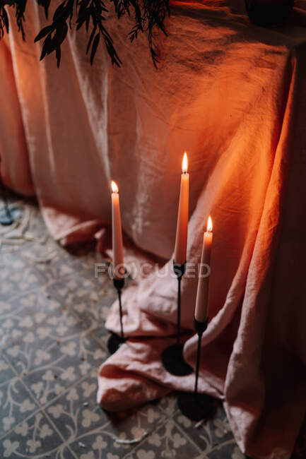 Сверху горящие восковые свечи на подсвечниках против складчатой ткани на декоративном кафельном полу в здании — стоковое фото