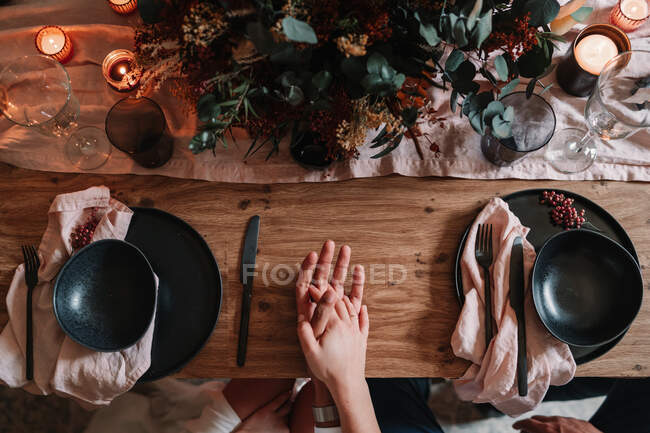 Top view of crop anonymous groom and bride holding hands on served table with flower decor and burning candles in restaurant — Stock Photo