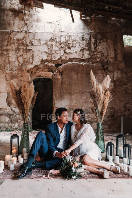 Full length young ethnic newlywed couple wearing posh wedding clothes lying on floor and looking at each other in grunge abandoned building room — Stock Photo